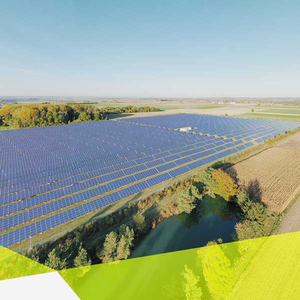 leichte Vogelperspektive auf einen Solarpark, der von links ins Bild ragt, umgeben von Feldern und Bäumen. Das obere Drittel ist Himmel ohne klare Wolkenbildung.