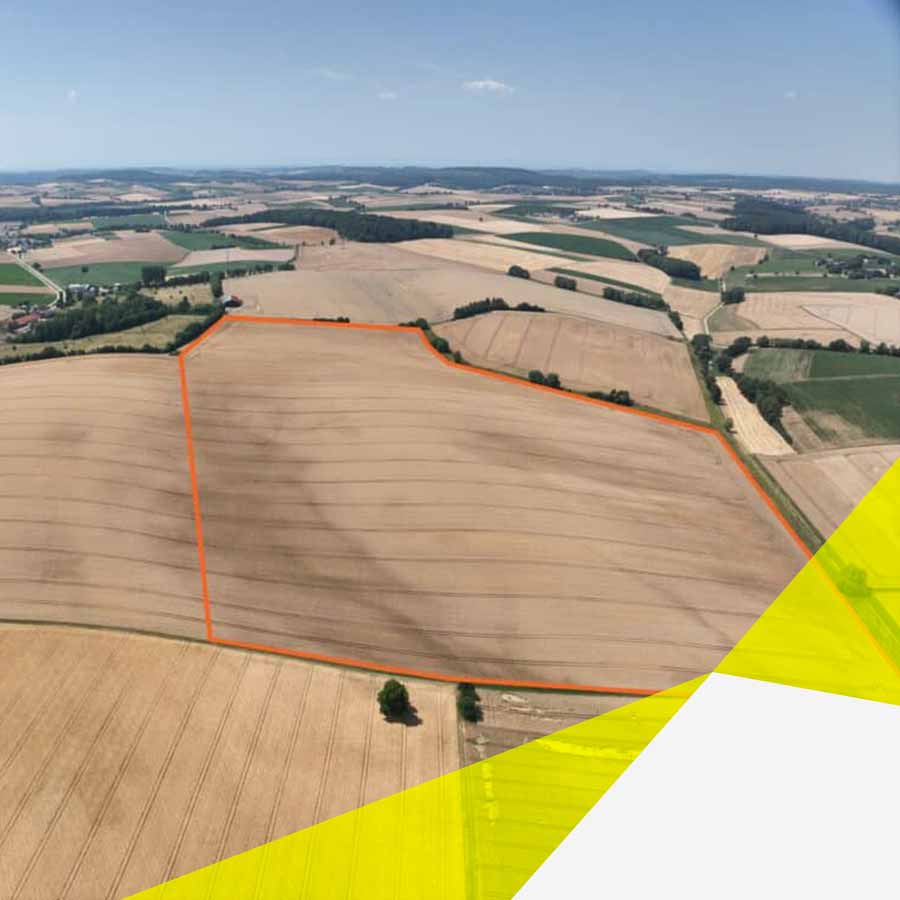 Es ist eine nicht sehr hohe Vogelperspektive. Am oberen Horizont ist Himmel, mit ganz wenigen kleinen Wolken zu erkennen. Neben vielen Feldern, ist in der Mitte ein ganz großes Feld, welches mit einer orangenen Linie markiert wurde. Die Linie kennzeichnet den zukünftigen Solarpark Buckeläcker.