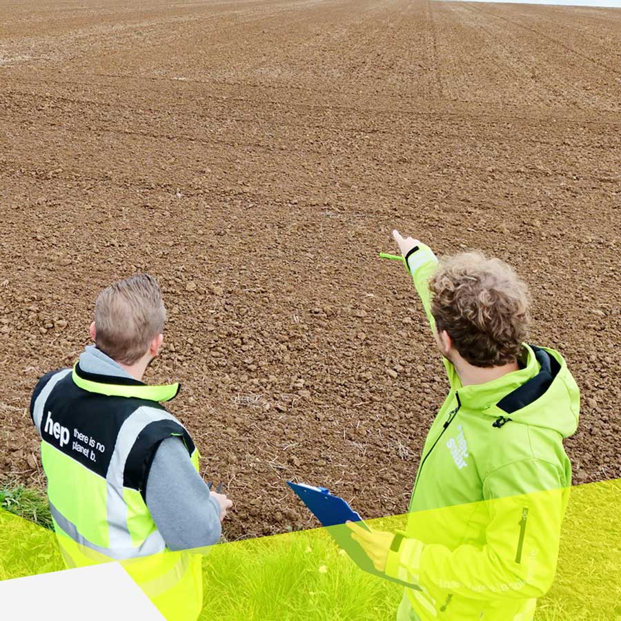 Zwei Personen stehe vorne und schauen auf ein braunes Feld. Der rechte hebt den rechten Arm und zeigt Richtung Feld.