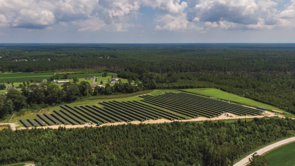 Eine Weitwinkelaufnahme eines Solarparks bestehend aus 35 Reihen mit Solapanels. Vor dem Park ist ein langer Sand Weg zusehen und drum herum eine Menge Bäume.