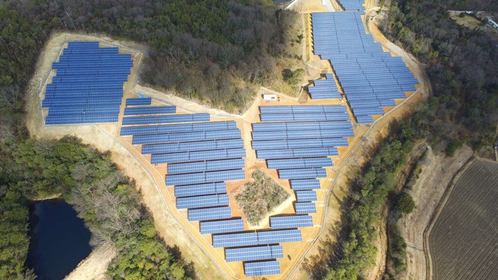 Ein Solarpark aus der Vogelsperspektive, Der Park hat eine Herzform und besteht aus ca. 30 Reihen. in der Mitte des Herzens ist eine kleine grüne Fläche zusehen, um den Park sind Bäume, links in der Ecke des Bild sieht man einen angrenzenden See.