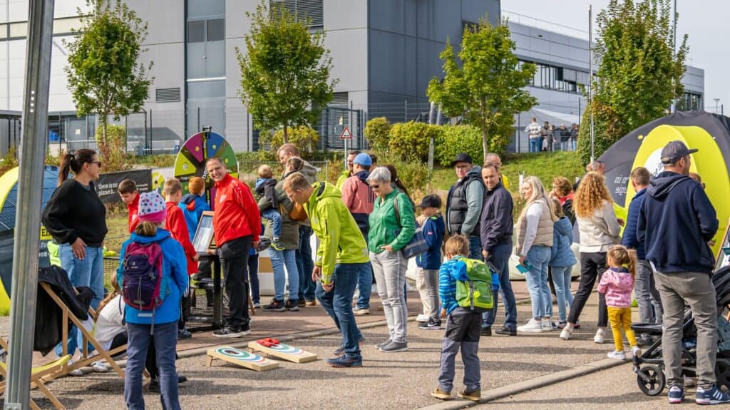 Eine Menge von Personen stehen Schlange um an einem Glücksrad zu drehen, dahinter ist ein schwarzes Zelt und ein dunkelgraues Gebäude zu sehen. Davor spielen Personen mit Sandsäcken, die in eine Zielscheibe auf den Boden geworfen werden müssen.