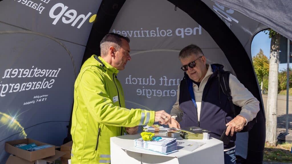 Zwei Personen stehen unter einem schwarzen bedrucktes Zelt. Die linke Personen trägt eine hellgrüne Jacke mit hep solar Logo und die rechte Person trägt eine Weste und Rucksack und schaut auf einen Stapel von Flyern, die mittig auf einen Stehtisch vor den beiden liegen.
