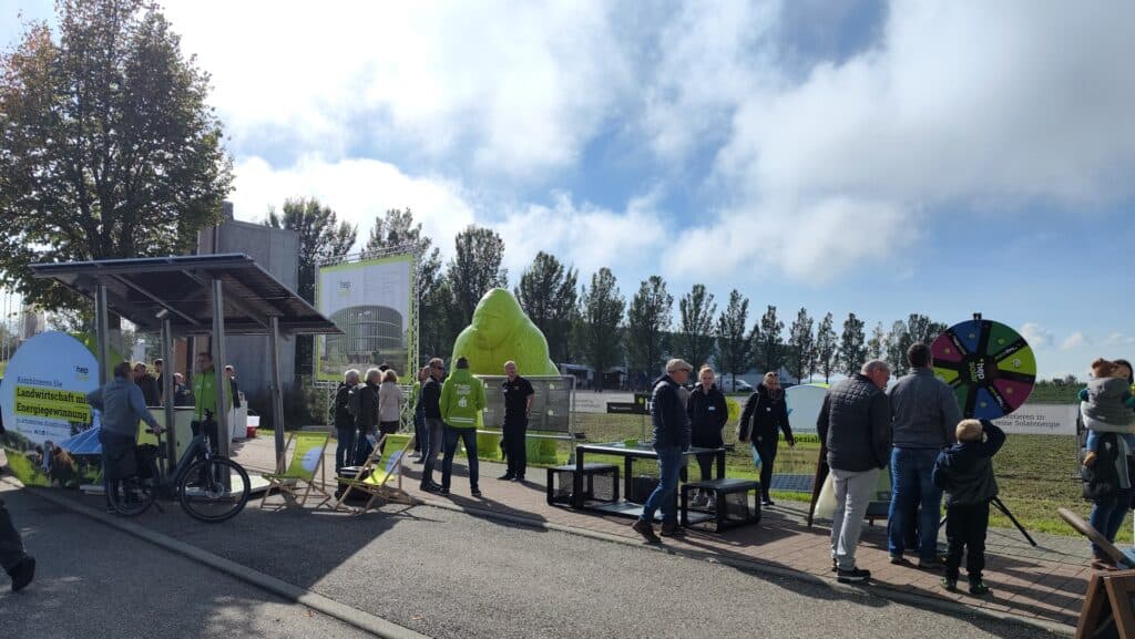Man sieht einige Menschen auf der Reihe verteilt, die entweder an einem Drehrad ihr Glück versuchen, an einem Tisch Kicker stehen, oder links vor einem schmalen Banner stehen. Im Hintergrund sieht man weitere Banner einen riesigen Gorilla und einen großen Industriebau, mit eine Reihe von Bäumen davor.