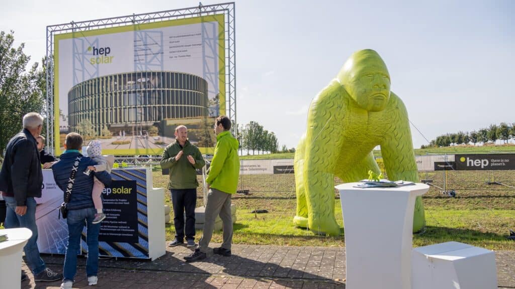 Im Hintergrund ist ein großes Feld zusehen, davor steht ein Bauschild mit hep solar Logo und einem runden Gebäude. Daneben ein riesiger Gorilla, mit Luft aufgepustet. Ganz vorne auf dem Weg stehen Personen und unterhalten sich.
