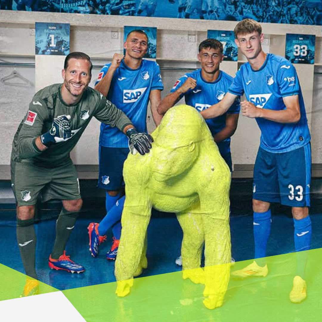 TSG Spieler und der Torwart stehen in der Umkleider in Ihren Spielerkleidung und jubeln. Sie stehen um einen neongrünen fast 1,30 Meter hohen Gorilla. Der Gorilla steht auf vier Beinen und schaut leicht nach links.