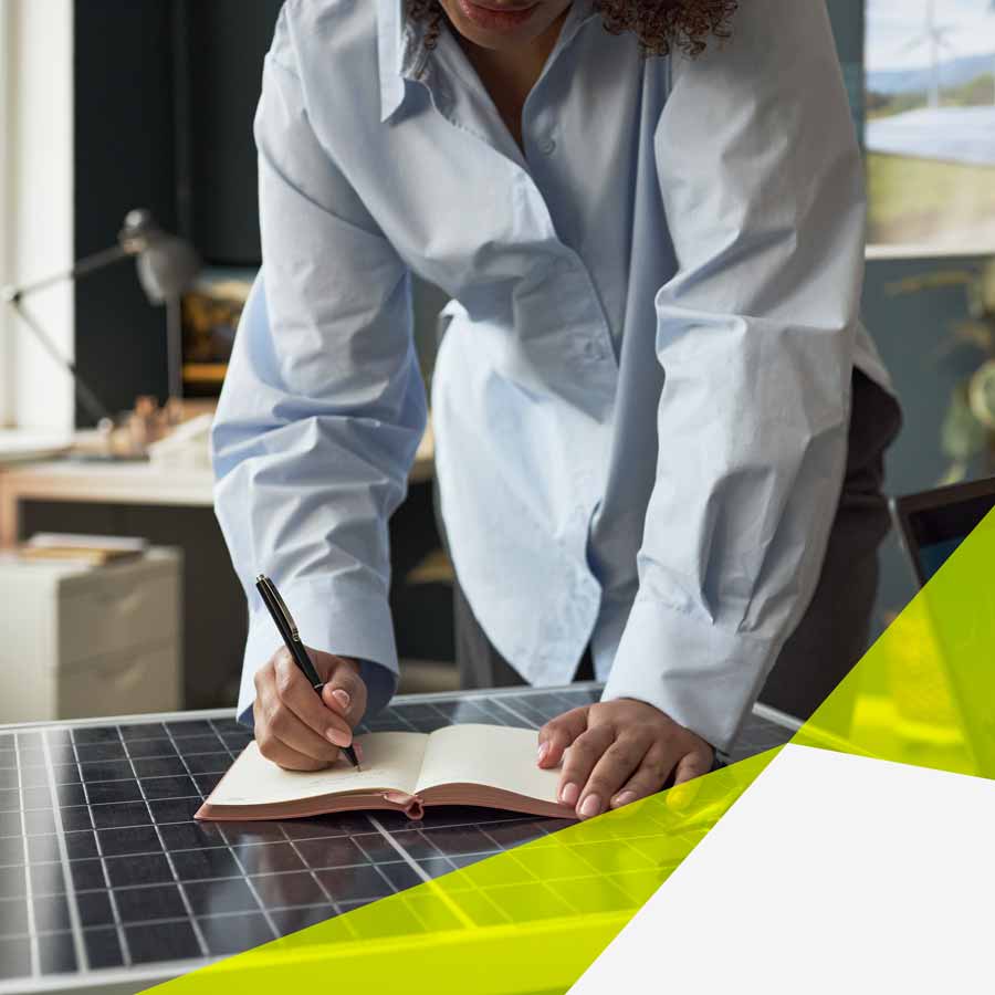 Eine Person stützt sich auf dem Tisch ab, mit gestreckten Armen und macht Notizen in einem kleinen Buch. Das Buch liegt auf einem Solar Panel. Im Hintergrund erkennt man einen Raum mit Schreibtisch, PC und Pflanzen.