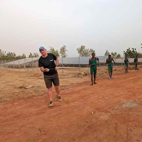 Matthias Hamann in Burkina Faso zieht ein dickes schwarzes Kabel, und wir von vier weiteren Personen hinter ihm, bilden sie eine Reihe und halten das Kabel. Im Hintergrund sind man eine Solaranlage. Die Erde ist sehr trocken und es stehen wenige Bäume hinter der Solaranlage.
