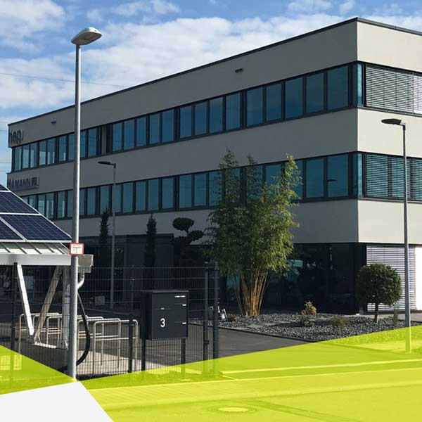 Rechteckiges Gebäude, mit drei Etage, pro Etage eine durchgehende Reihe Fensterscheiben, links hinten ist ein hep Logo und ein Hamann Logo zu sehen.