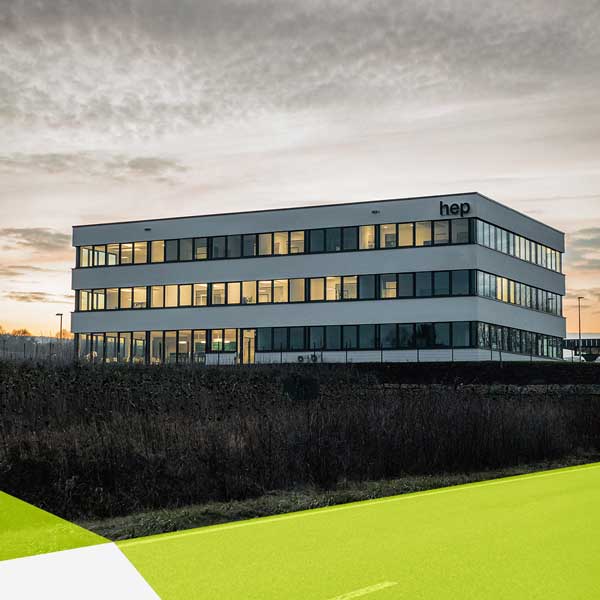 Ein Gebäude bei Sonnenuntergang. Die Sonne spiegelt sich in den Fenstern wieder. Das weiße, dreistöckige Gebäude hat oben rechts ein hep Logo.