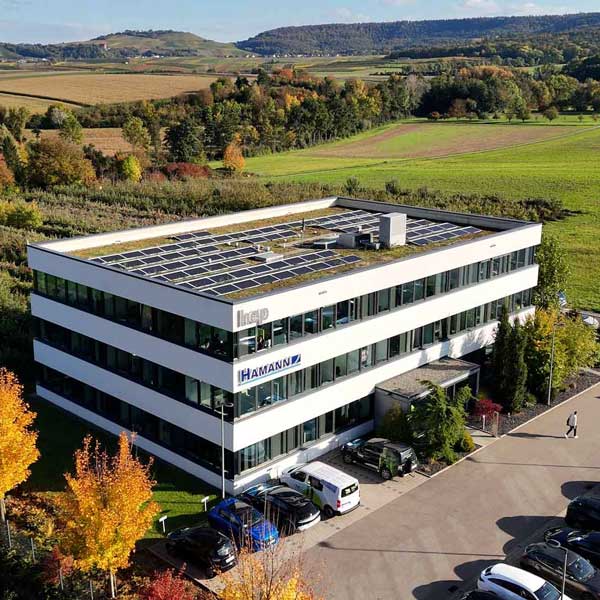 Aus der Vogelperspektive sieht man eine hügelige Landschaft mit Feldern, Bäume mit grünen, gelben und roten Baumkronen und in der Mitte ein Gebäude mit Solarpanels auf dem Dach. Das Haus ist rechteckig und optische geschichtet eine weiße Fassade, dann ein Ring aus Fenstern, dann wieder ein Ring weiße Fassade und dann wieder Fenster. Insgesamt dreistöckig und es sind zwei Logos außen angebracht, einmal hep und einmal Hamann.